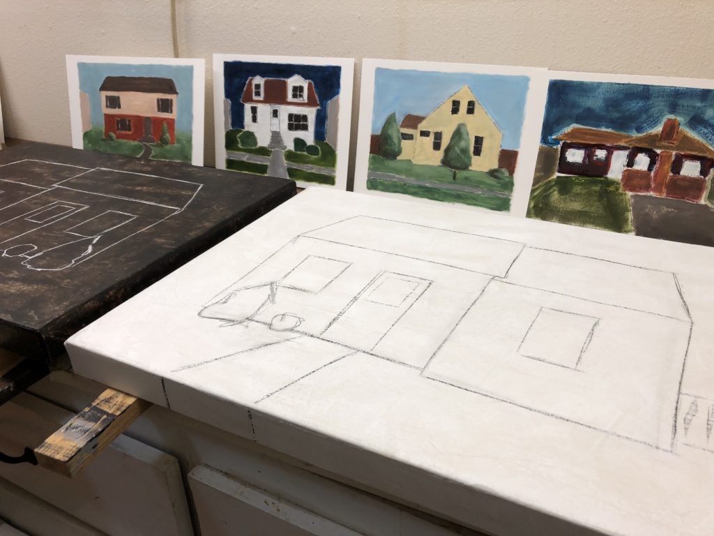 Black and white paintings of houses in progress on a work table with small paintings of houses in background against wall. 