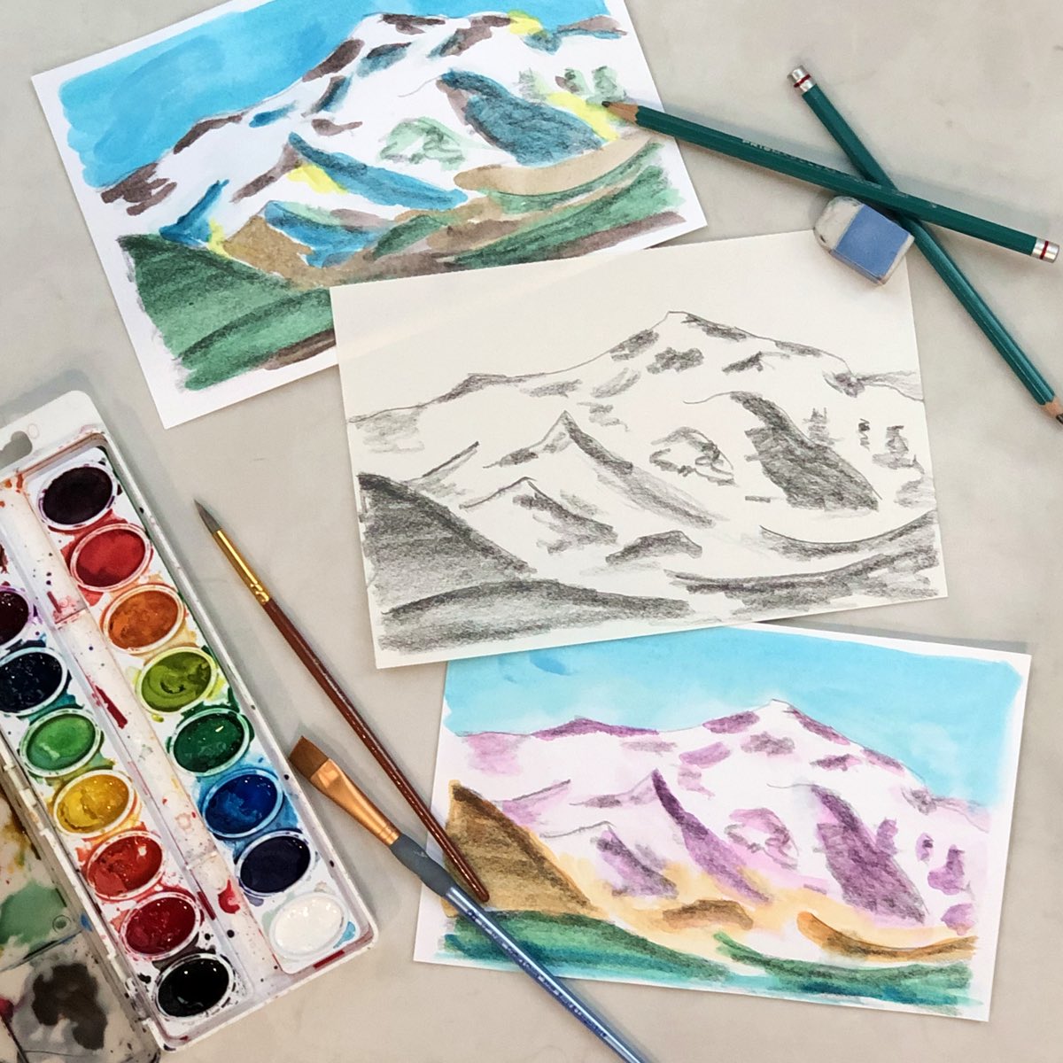 Pencil drawing and watercolor painting of mountain landscape with drawing pencils paint brushes and paint set.